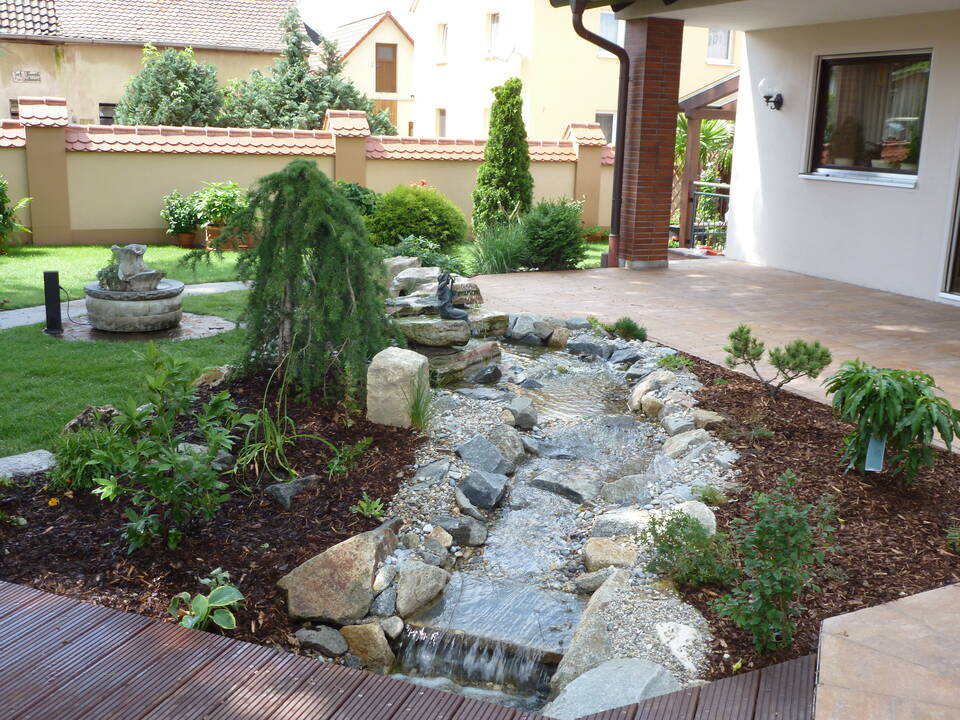 Garten-Bachlauf fließt von einem Quellstein mit Bronze-Figur zu einer Holzterrasse, unter der er mit einem kleinen Wasserfall verschwindet.