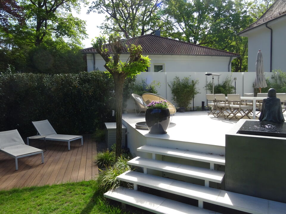 Rechts eine weiße, erhöhte Terrasse mit Treppe, Wasserspiel und Gartentisch, links eine ebenerdige Holzterrasse mit 2 Liegestühlen. Beispiele für Terrassenbau in Nürnberg.
