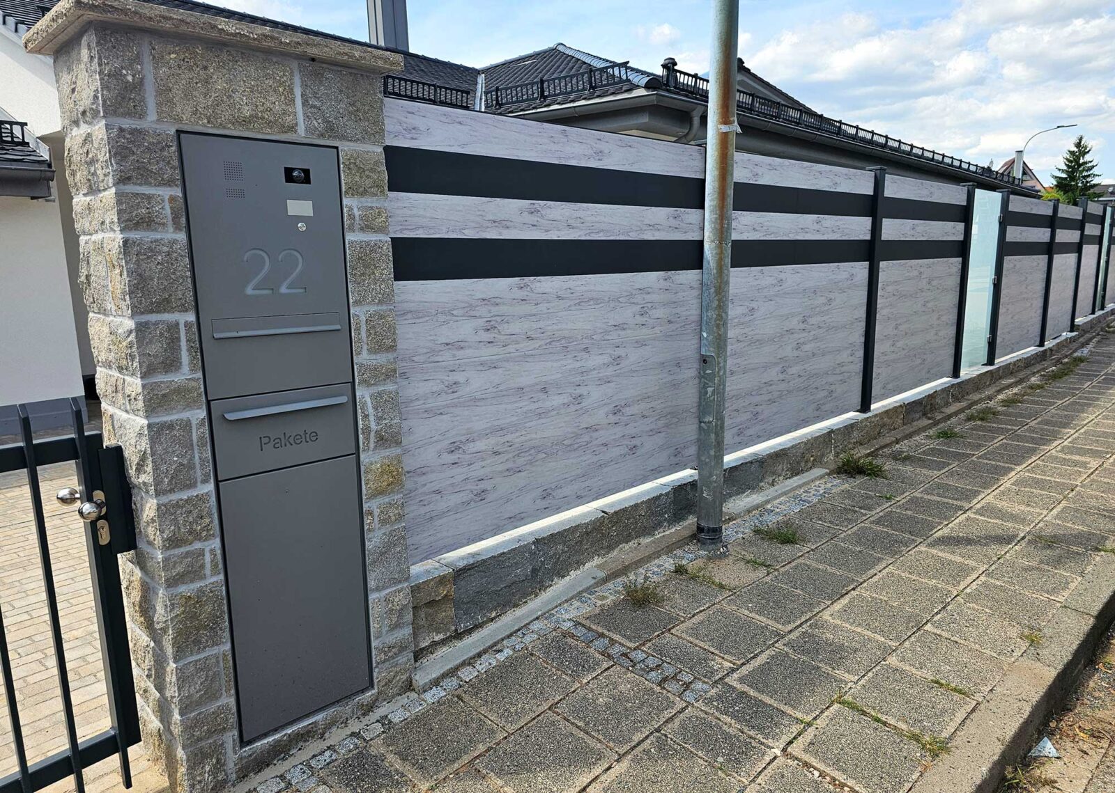 Sichtschutz aus WPC-Platten mit Holzoptik und grauen Streifen, unterbrochen von satinierten Glasscheiben als Gartenzaun. Links grau lackiertes Stahl-Gartentor sowie aus Naturstein gemauerter Briefkasten mit Paketbox, Klingel, beleuchteter Hausnummer und Kamera.
