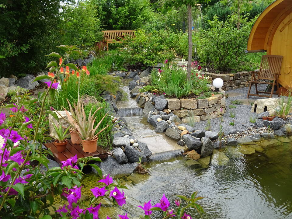 Neben einer Faßsauna fließt ein Garten-Bachlauf über 2 Wasserfälle in einen Gartenteich.