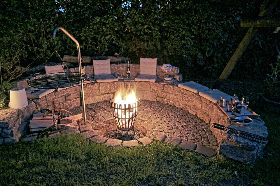 Gemauerte Feuerstelle im Garten bei Ansbach mit brennendem Feuerkorb, weggeschwenktem Grillrost und grauen Polstern am Abend