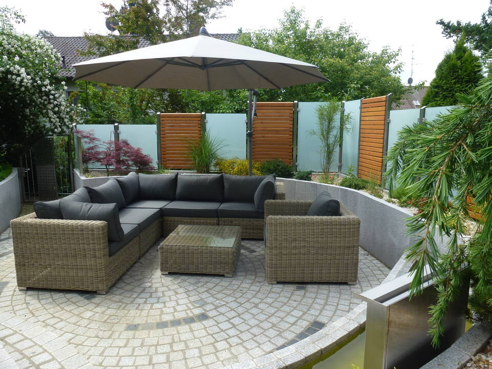 Garten-Lounge mit Korb-Gartensofa, weißem Sonnenschirm und Sichtschutz im Hintergrund. Der Sichtschutz kombiniert Edelstahl-Pfosten mit Elementen aus Milchglas und Holz. Links eine Gartentür aus Edelstahl.