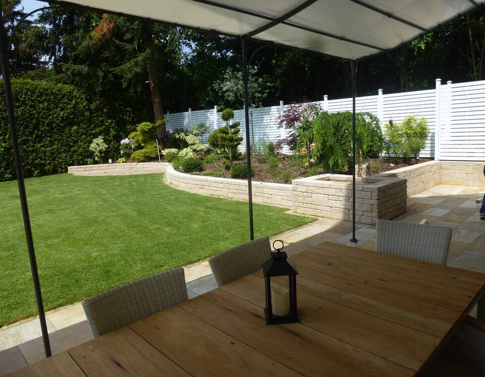Überdachte Terrasse mit Gartentisch, dahinter gemauertes Hochbeet und weißer Sichtschutz.