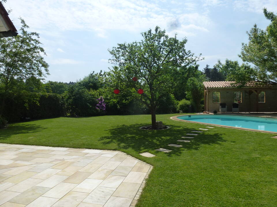 Blick von der Terrasse über den Rasen zu einem Pool mit überdachter Mauer als Sonnenschutz.