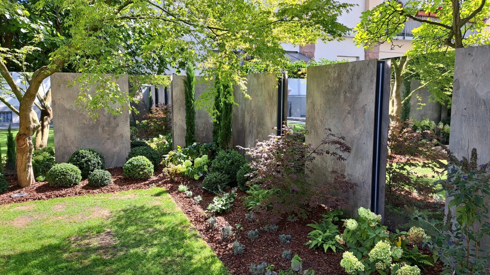 Sichtschutz in der Ecke eines Gartens: Spiegel wechseln sich mit Verbundplatten in Beton-Optik ab.