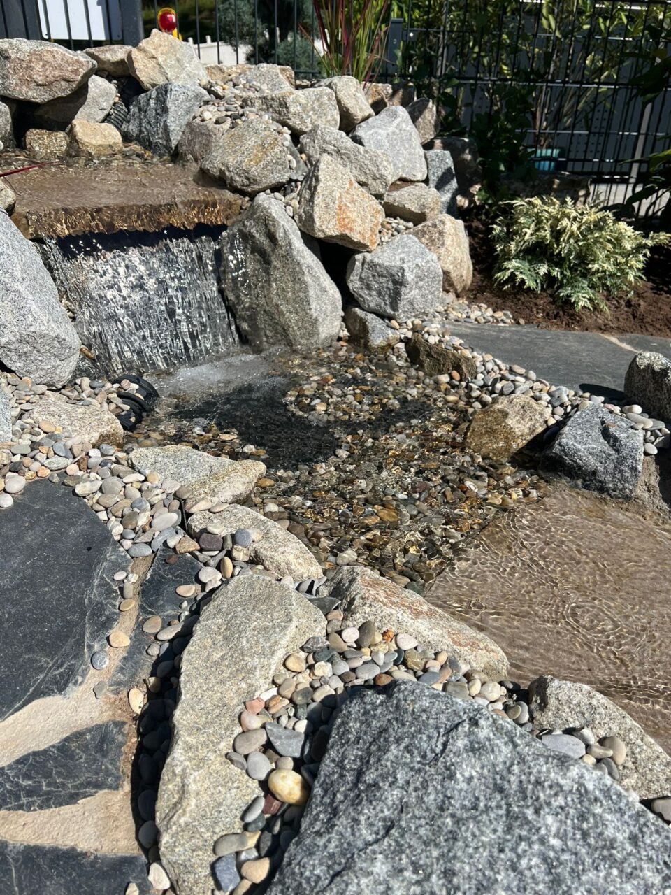 Ein von Natursteinen umrahmter Bachlauf im Garten entspringt aus einem künstlichen Hügel und fließt über einen kleinen Wasserfall.