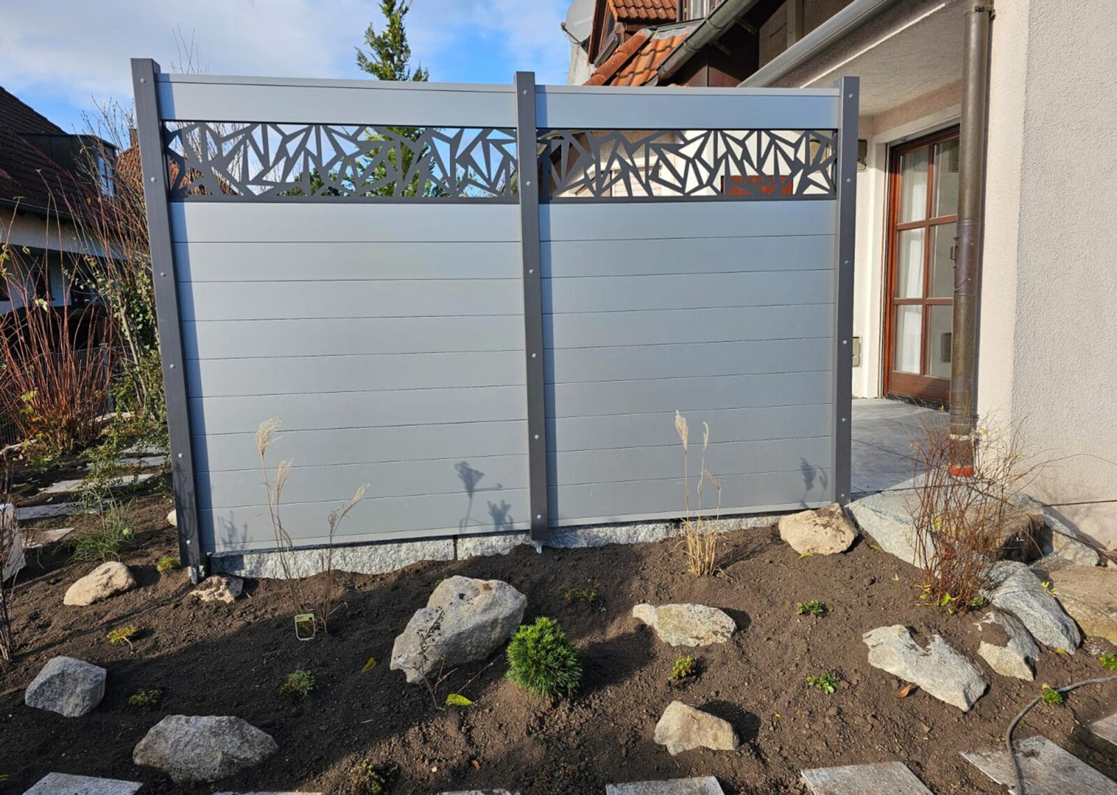 Hellgrauer Terrassen-Sichtschutz aus grau lackierten Stahlpfosten, WPC-Platten und einem gemusterten Element.