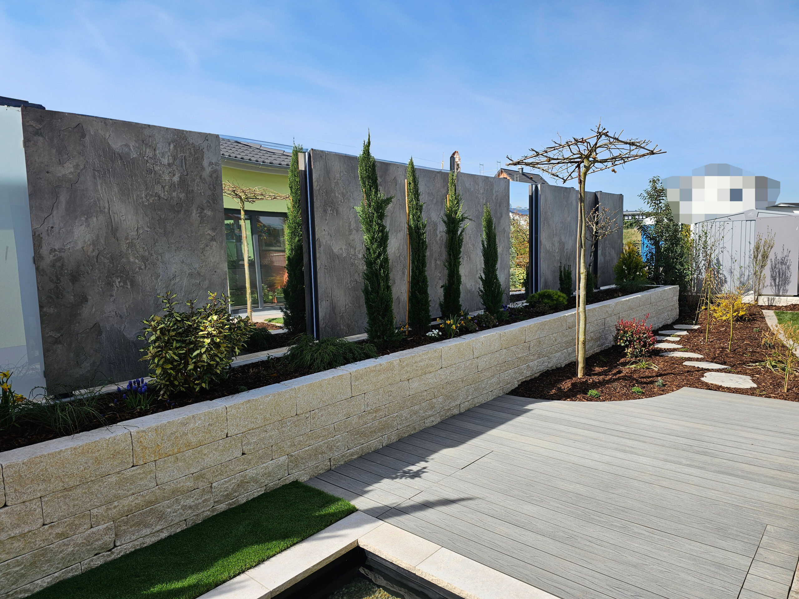 Schattenplatz-Terrasse mit Schirmplatane (noch ohne Blätter) vor einem Beet mit Trittsteinen. Links Sichtschutz-Wand aus WPS-Platten und Spiegeln hinter einem Hochbeet aus weißem Naturstein.