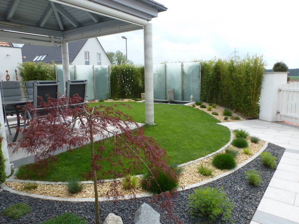 Bambus-Hecke als Sichtschutz und Gartenzaun. Elemente aus Milchglas-Scheiben und Edelstahl-Zaunpfählen lockern die Hecke auf. Daneben eine weiße Holz-Gartentür, die von einem massiv gemauerten Zaunpfahl gehalten wird.