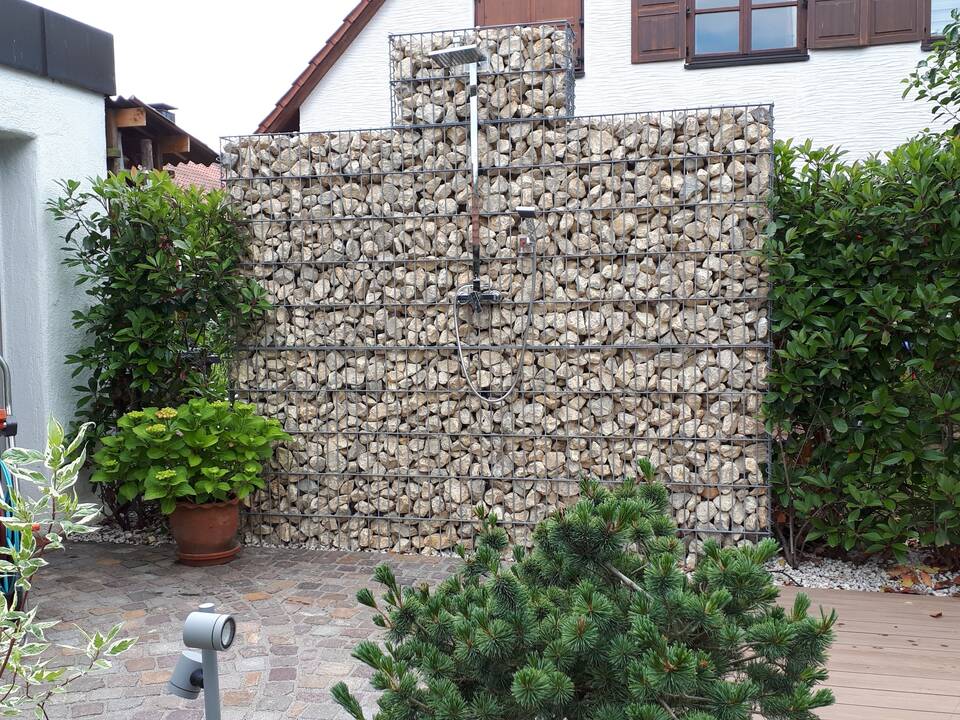 Eine große Gabione dient sowohl als Sichtschutz als auch als Befestigungsmöglichkeit für eine Gartendusche. Daneben Kirschlorbeer-Hecke.