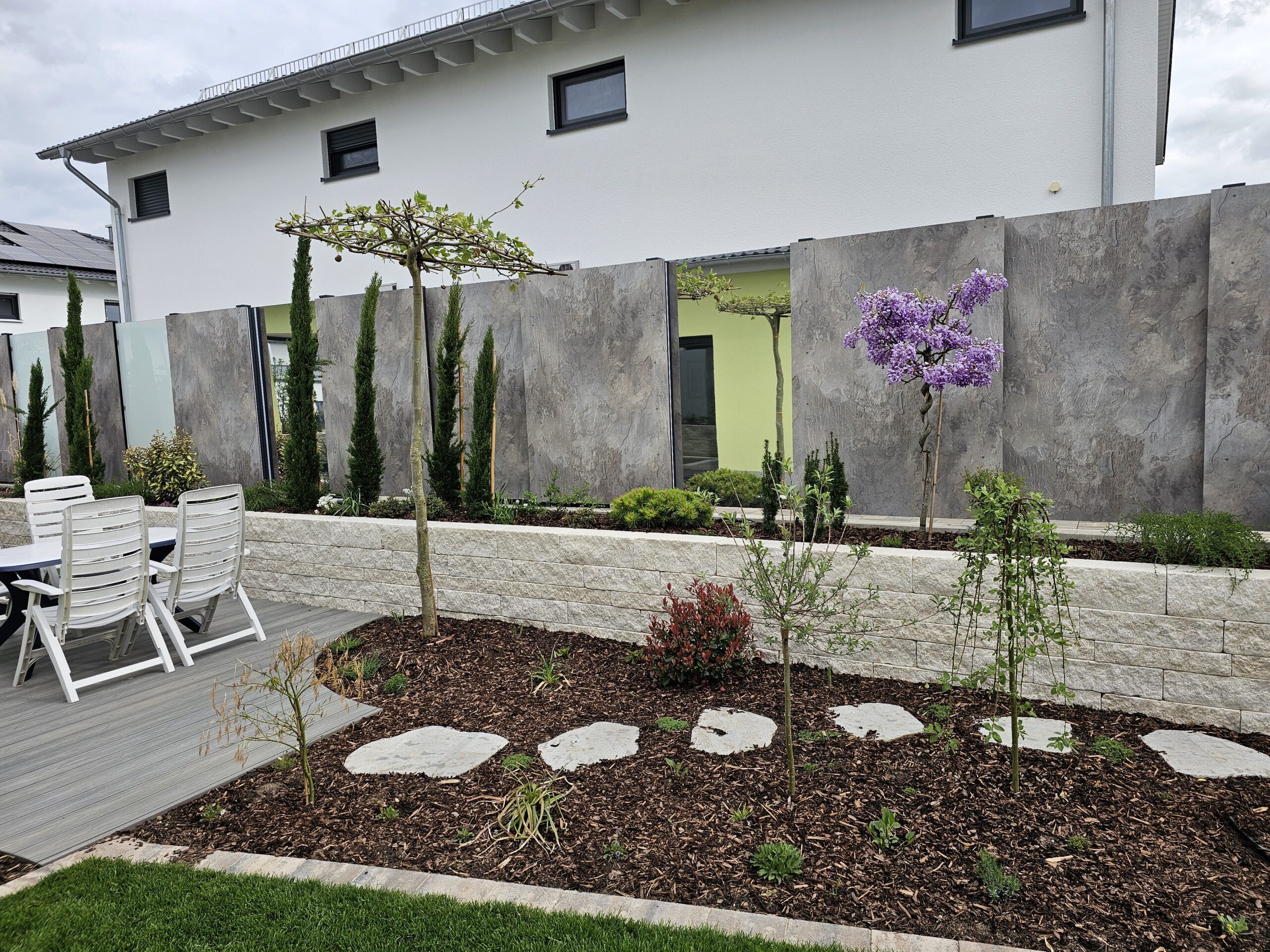 Beet mit Rindenmulch und weißen Naturstein-Trittsteinen in einem Garten in Ansbach. Dahinter Hochbeet und Sichtschutz, links Sitzecke aus WPC-Dielen mit einer Schirmplatane als Schattenspender.