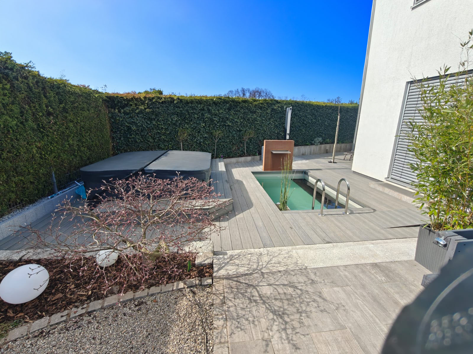 Whirlpool auf erhöhtem Podest, daneben Tauchbecken mit Edelstahl-Poolleiter und Unterwasser-Sitzbank. Beides umgeben von einer Holzterrasse. Im Hintergrund Cortenstahl-Wasserschütte und Solar-Gartendusche. Die Sauna ist neben dem Gebäude um die Ecke.